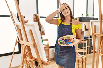 Young hispanic woman at art classroom stressed and frustrated with hand on head, surprised and...