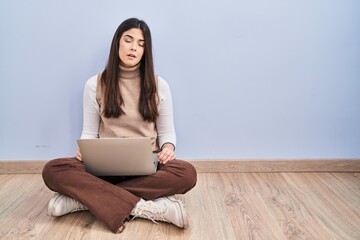 Young brunette woman working using computer laptop sitting on the floor looking sleepy and tired, exhausted for fatigue and hangover, lazy eyes in the morning.