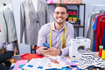Young hispanic man tailor smiling confident using smartphone at tailor shop