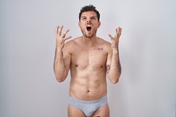 Fototapeta premium Young hispanic man standing shirtless wearing underware crazy and mad shouting and yelling with aggressive expression and arms raised. frustration concept.