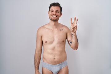 Fototapeta premium Young hispanic man standing shirtless wearing underware showing and pointing up with fingers number three while smiling confident and happy.