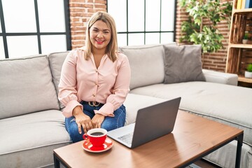 Young hispanic woman using laptop sitting on the sofa at home winking looking at the camera with...