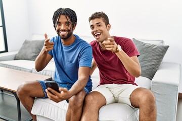 Young hispanic men using smartphone sitting on the sofa at home pointing fingers to camera with happy and funny face. good energy and vibes.