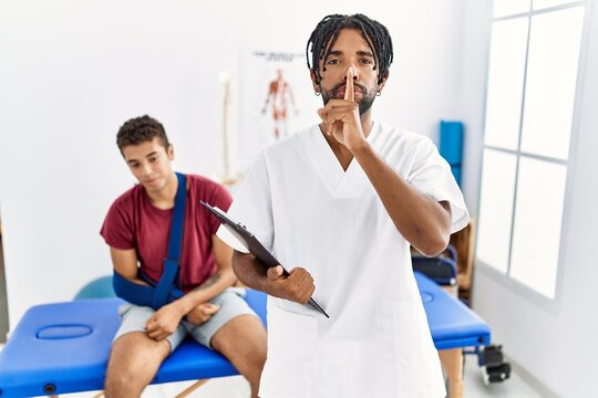 Young hispanic man working at pain recovery clinic with a man with broken arm asking to be quiet with finger on lips. silence and secret concept.