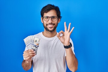 Naklejka premium Handsome latin man holding dollars banknotes doing ok sign with fingers, smiling friendly gesturing excellent symbol