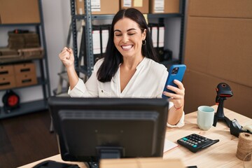 Young hispanic woman working at small business ecommerce with smartphone screaming proud, celebrating victory and success very excited with raised arm