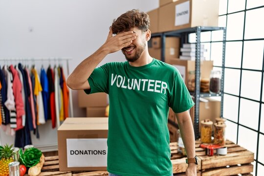 Young arab man wearing volunteer t shirt at donations stand smiling and laughing with hand on face covering eyes for surprise. blind concept.