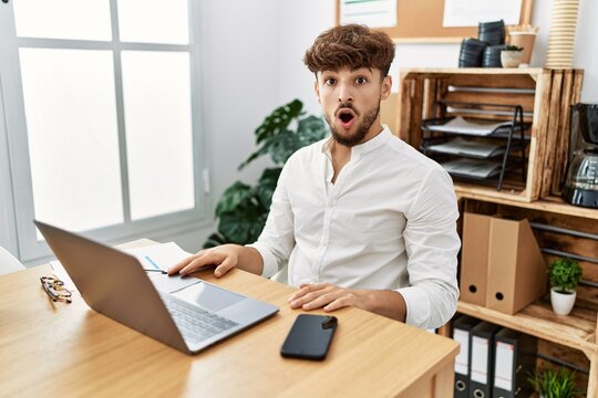 Young arab man working using computer laptop at the office scared and amazed with open mouth for surprise, disbelief face