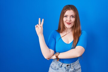 Redhead woman standing over blue background smiling with happy face winking at the camera doing victory sign with fingers. number two.