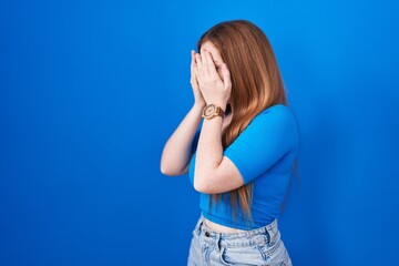 Redhead woman standing over blue background with sad expression covering face with hands while...