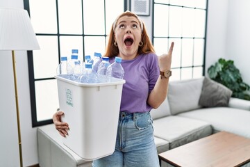 Young redhead woman holding recycling wastebasket with plastic bottles amazed and surprised looking up and pointing with fingers and raised arms.