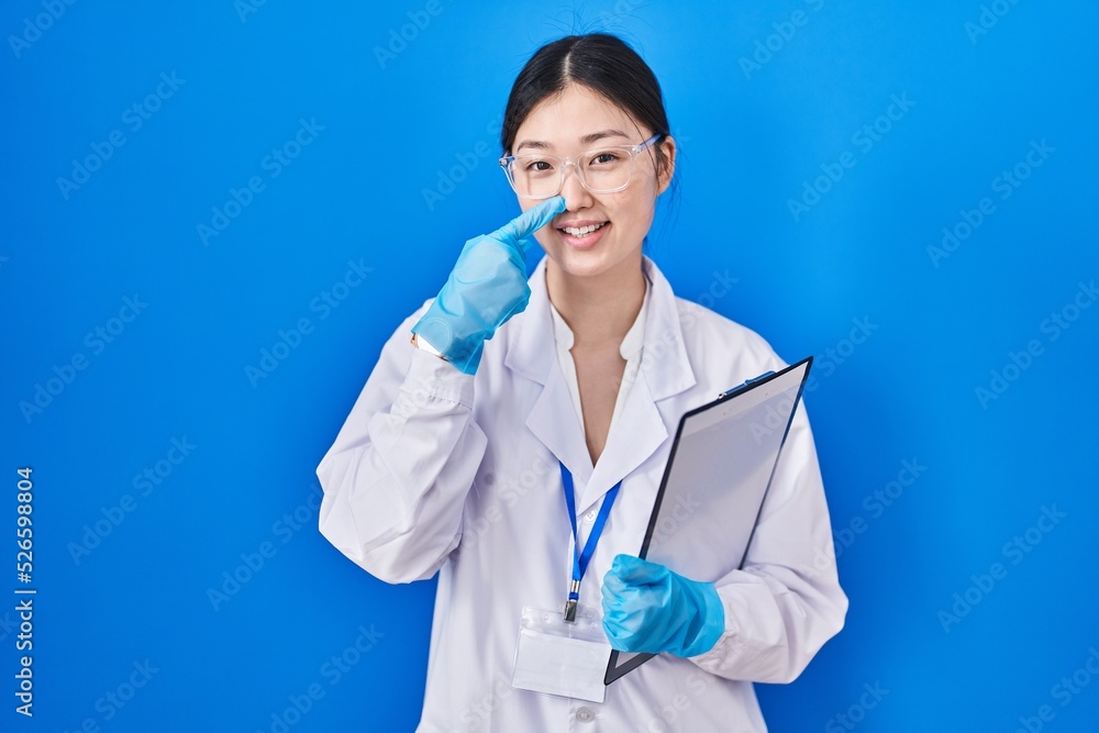 Poster chinese young woman working at scientist laboratory pointing with hand finger to face and nose, smil