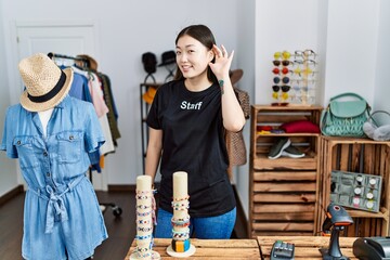 Young asian woman working as manager at retail boutique smiling with hand over ear listening an hearing to rumor or gossip. deafness concept.