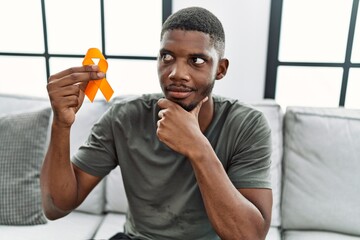 Young african american man holding awareness orange ribbon sitting on the sofa serious face...