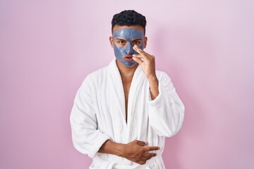 Young hispanic man wearing beauty face mask and bath robe mouth and lips shut as zip with fingers. secret and silent, taboo talking