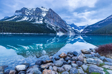 Clear blue lake with snowy mountain reflections and colorful river rock foreground. 