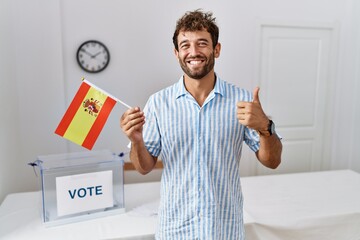 Young handsome man at political campaign election holding spain flag smiling happy and positive, thumb up doing excellent and approval sign