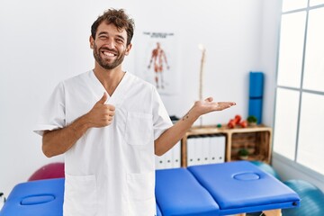 Young handsome physiotherapist man working at pain recovery clinic showing palm hand and doing ok gesture with thumbs up, smiling happy and cheerful