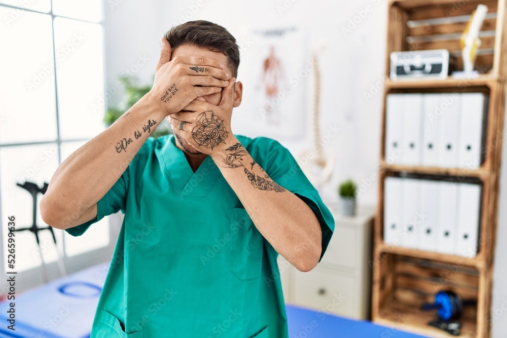 Canvas Prints Young physiotherapist man working at pain recovery clinic covering eyes and mouth with hands, surprised and shocked. hiding emotion