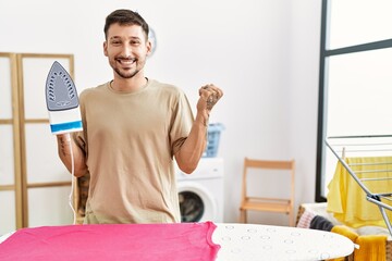 Young handsome man ironing clothes at home screaming proud, celebrating victory and success very excited with raised arm