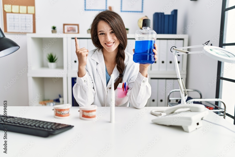 Poster young dentist woman holding mouthwash for fresh breath smiling with an idea or question pointing fin