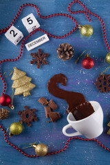 Christmas mood. Top view on small white tea cup with spilled out hot chocolate on abstract blue background. Holiday decorations and wooden cube calendar 25 December date. Top view, flat lay. Vertical