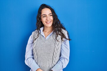 Fototapeta premium Young brunette woman standing over blue background looking away to side with smile on face, natural expression. laughing confident.