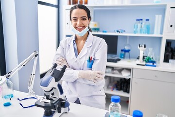 Young hispanic woman wearing scientist uniform standing with arms crossed gesture at laboratory