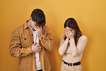 Young hispanic couple standing over yellow background with sad expression covering face with hands...