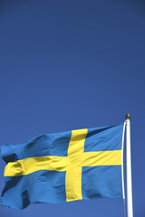 Die Nationalfahne von Schweden in den Faben blau und gelb weht im Wind an einem Fahnenmast. Die Krone ist die Währung von Schweden. Als Hintergrund dient der blaue Himmel.