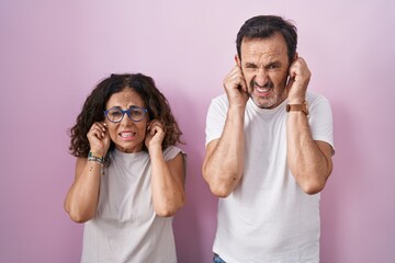 Middle age hispanic couple together over pink background covering ears with fingers with annoyed expression for the noise of loud music. deaf concept.