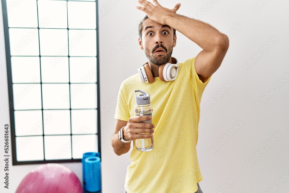 Wall mural young hispanic man wearing sportswear and drinking water at the gym surprised with hand on head for 