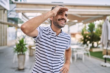 Young hispanic man with beard outdoors at the city very happy and smiling looking far away with hand over head. searching concept.