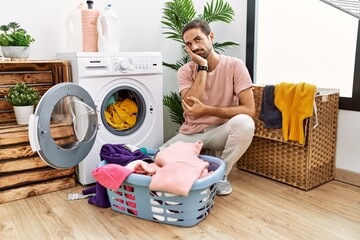 Young hispanic man putting dirty laundry into washing machine thinking looking tired and bored with depression problems with crossed arms.