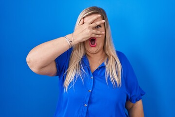 Caucasian plus size woman standing over blue background peeking in shock covering face and eyes...