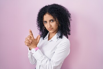 Hispanic woman with curly hair standing over pink background holding symbolic gun with hand gesture, playing killing shooting weapons, angry face