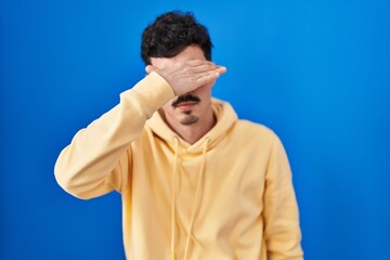 Hispanic man standing over blue background covering eyes with hand, looking serious and sad....