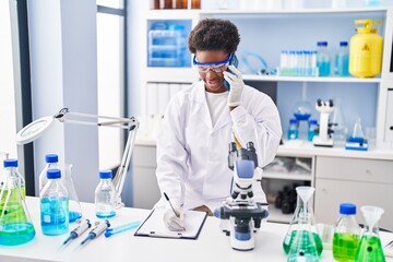 African american woman wearing scientist uniform talking on the smartphone write on clipboard at...