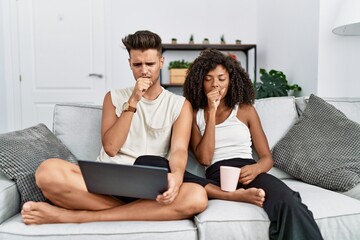 Young interracial couple using laptop at home sitting on the sofa feeling unwell and coughing as symptom for cold or bronchitis. health care concept.