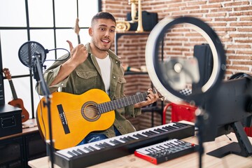 Young hispanic man doing online guitar tutorial pointing thumb up to the side smiling happy with...
