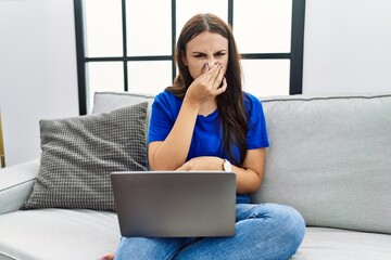 Young brunette woman using laptop at home smelling something stinky and disgusting, intolerable...