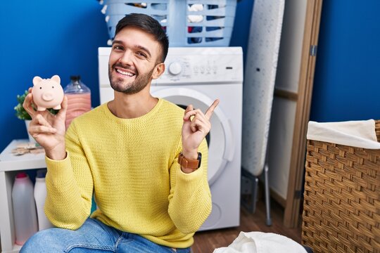 Hispanic man doing laundry holding piggy bank smiling happy pointing with hand and finger to the side