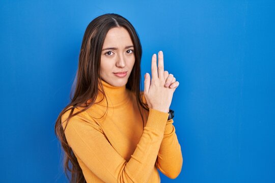 Young brunette woman standing over blue background holding symbolic gun with hand gesture, playing killing shooting weapons, angry face