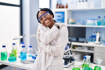 African american woman working at scientist laboratory hugging oneself happy and positive, smiling...