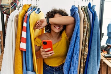Young hispanic woman searching clothes on clothing rack using smartphone smiling cheerful playing peek a boo with hands showing face. surprised and exited