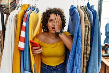 Young hispanic woman searching clothes on clothing rack using smartphone yawning tired covering...