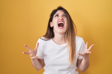 Young brunette woman standing over yellow background crazy and mad shouting and yelling with aggressive expression and arms raised. frustration concept.