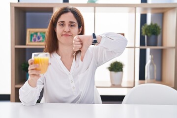 Brunette woman drinking glass of orange juice looking unhappy and angry showing rejection and negative with thumbs down gesture. bad expression.