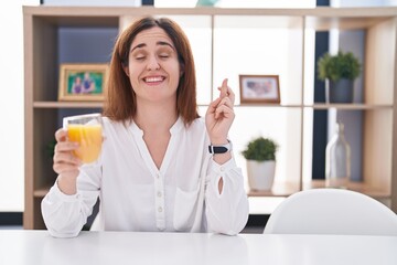 Brunette woman drinking glass of orange juice gesturing finger crossed smiling with hope and eyes closed. luck and superstitious concept.