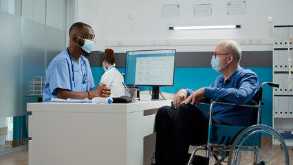 Male nurse doing checkup consultation with elderly man in wheelchair, having appointment in cabinet. Medical assistant consulting patient with chronic disability during covid 19 pandemic.